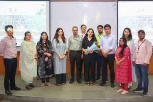 Group of people on a stage posing for a photo, some holding flowers and a trophy.