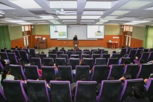 Speaker at a wooden podium delivering a lecture to an auditorium filled with students seated in purple chairs
