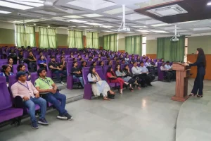 Speaker at a podium giving a lecture to an audience in a lecture hall with purple upholstered seating and green curtains.