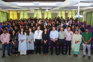 Large group of professionals posing for a group photo in a bright auditorium with green curtains and purple seating in the background, everyone looking at the camera.
