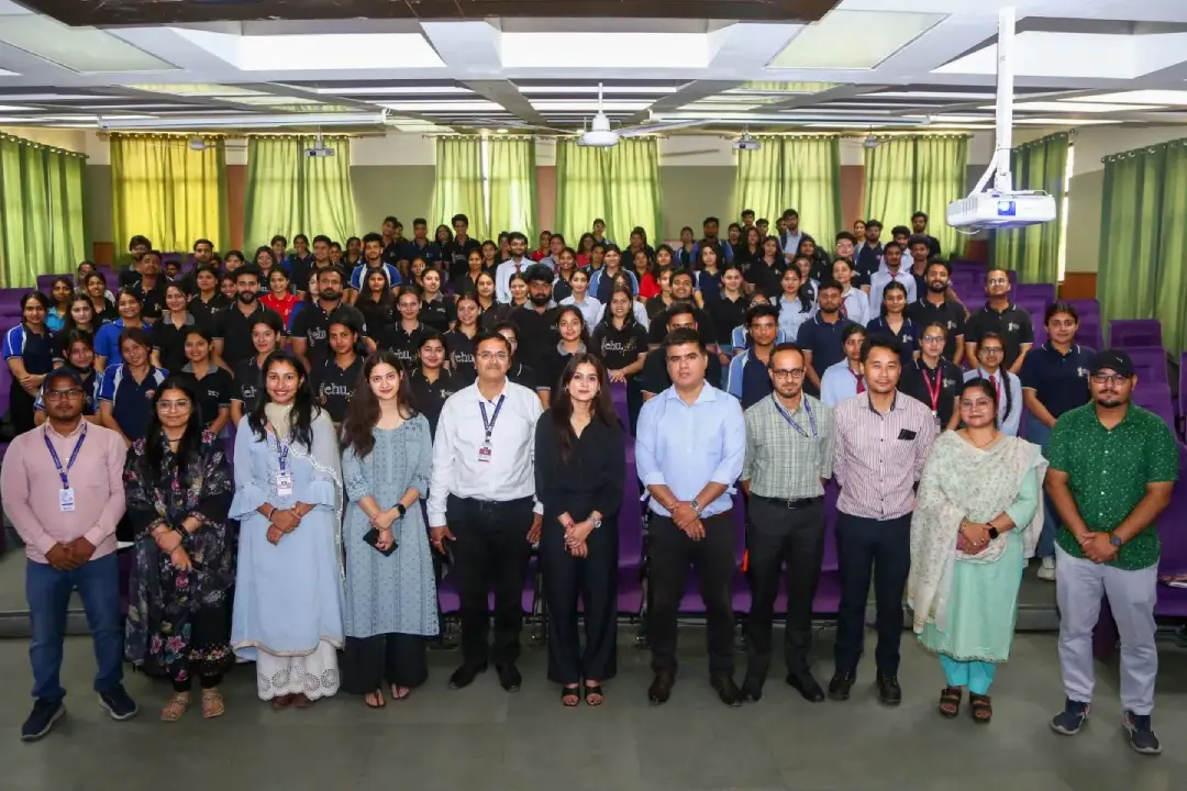 Large group of professionals posing for a group photo in a bright auditorium with green curtains and purple seating in the background, everyone looking at the camera.