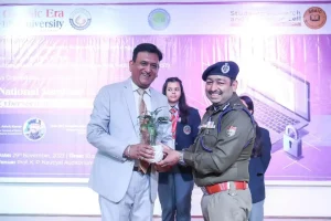Man in a light gray suit hands a small potted plant to a smiling police officer on stage during a ceremony, with a purple banner backdrop and a student in uniform in the background.