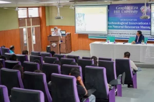 Speaker at a lectern presenting to an audience in a university lecture hall with a projector screen in use at the front.