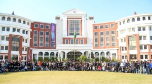Large group of students posing for a class photo in front of a red-brick university building with a central white entrance and flagpole.