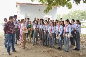 Group of students in blue shirts and red-striped ties listening to two instructors outdoors, briefing under a tree.