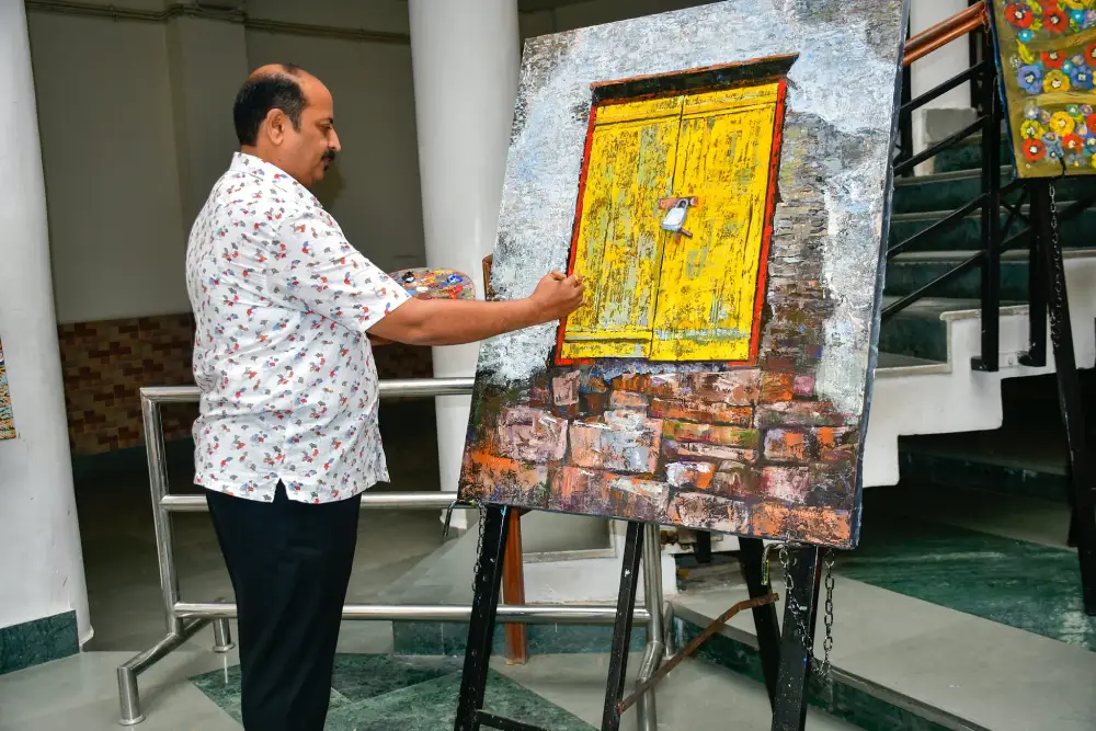 Man in a white floral shirt paints a large canvas on an easel, depicting a bright yellow door set in a brick wall.