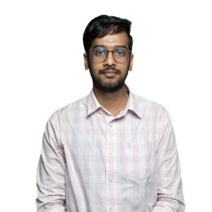 Portrait of a man with dark hair and beard, wearing round glasses and a pale pink plaid shirt against a white background.