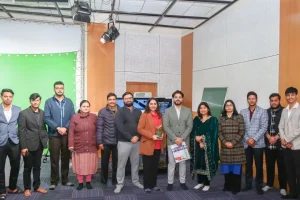 Group of people posing in a studio beside a green screen, some holding papers and a small plant in the center.