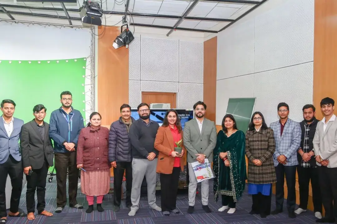 Group of people posing in a studio beside a green screen, some holding papers and a small plant in the center.