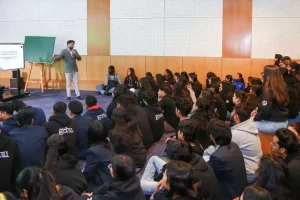 Presenter at the front speaking to a large group of students seated on the floor in a classroom. (informative)