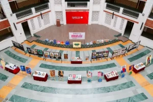 Wide shot of a circular, multi-tiered atrium hosting an annual art exhibition; art boards and tables display colorful artwork around a central banner.