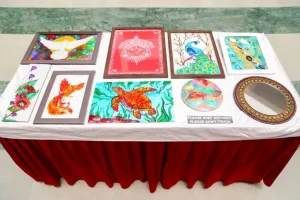 Table display of colorful framed artworks and a round decorative mirror on a white-clothed surface with red fabric beneath.