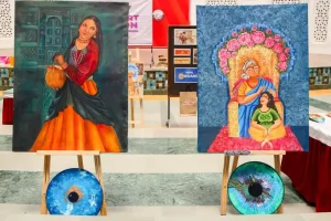 Two vibrant paintings on wooden easels in a gallery—left painting shows a woman in traditional dress holding a basket; right painting depicts an elderly woman blessing a young girl under a floral arch, with colorful circular artworks on the floor nearby.