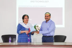 Woman in blue traditional attire receives a bouquet from a man in a light blue shirt at a formal event in front of a projection screen.