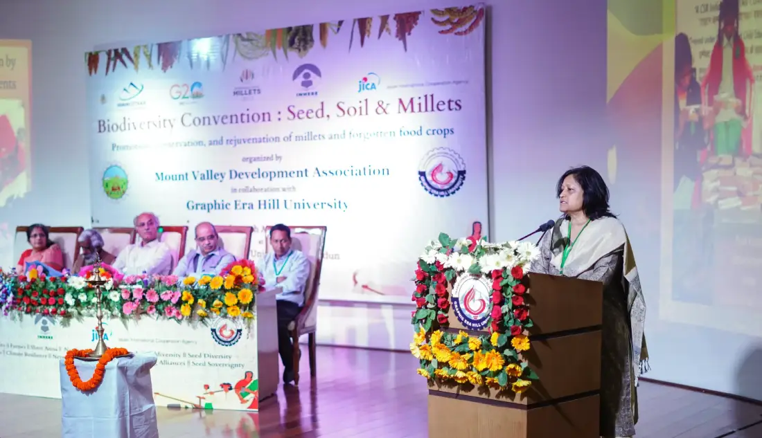 Speaker at a flower-decorated podium addressing a panel on stage; banner reads Biodiversity Convention: Seed, Soil & Millets with panelists seated behind.