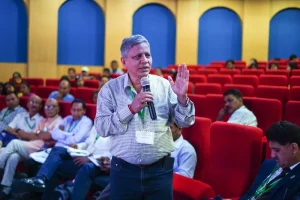 Man with microphone speaking to an audience in a modern auditorium with red seats and yellow walls, wearing a checkered shirt and conference badge.