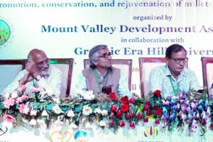 Panel of three men seated at a conference dais with flower arrangements, backdrop reads 'Mount Valley Development Association' and 'Graphic Era Hill University'