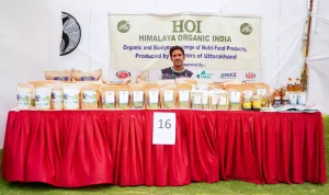 Vendor at a HOI Himalaya Organic India booth with many packaged products on a red-draped table; banner behind reads HOI Himalaya Organic India.