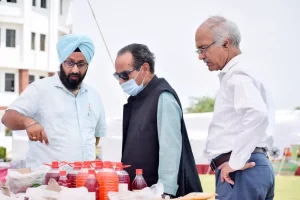 Three men discuss red beverages laid out on a table at an outdoor event; one wears a blue turban and glasses, another wears a mask and vest, the third observes from the right.