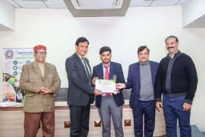 Five men in a room pose for a certificate award; the central young man in a suit and red tie accepts a certificate from a man in a dark suit, others nearby applaud.
