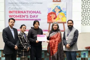 Five people stand on a stage as a certificate is handed to a young recipient during an International Women's Day event, with a colorful banner in the background.