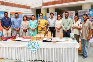 Group of people standing behind a display table with books and crafts at an indoor event; banners visible in the background.