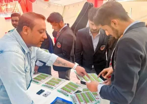 Man in a denim jacket hands out green pamphlets to suited young men at a booth.