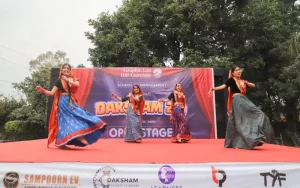 Four girls in colorful traditional outfits perform a dance on a red stage during a school event behind them a banner reads 'DAKSHAM 2'.