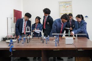 Group of students in uniform blazers and striped ties collaborating at a science lab bench with metal experimental rigs and instruments on display behind them.