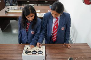 Two schoolgirls in blue uniforms with red striped ties stand at a wooden lab bench, adjusting controls on a vintage electrical training panel on a classroom table.
