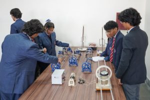 Group of students in blue uniforms and red striped ties gather around a lab table, studying various mechanical models and gears.
