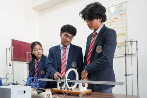 Three students in uniforms work together on a classroom mechanical model with blue gears and a belt-driven setup on a lab table.