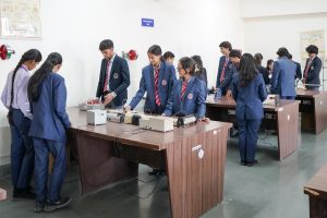 Students in blue uniforms and red ties work at lab benches with electrical/mechanical equipment in a mechanics lab.