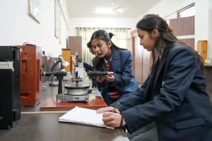 Two students in a school lab examine a precision instrument on the bench, one taking notes in a notebook.