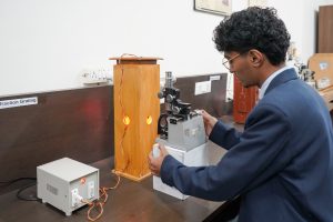 Researcher in a blue blazer works at a lab bench, adjusting a mechanical instrument mounted on a wooden frame with glowing orange components and wires nearby.