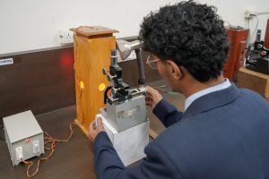 A person in a dark blue lab coat adjusts a metallurgical microscope connected to a wooden stand with glowing orange chambers in a laboratory setting.