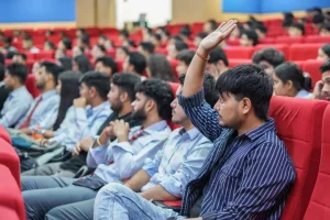Student in a blue-striped shirt raises hand during a lecture in a red-seat auditorium.