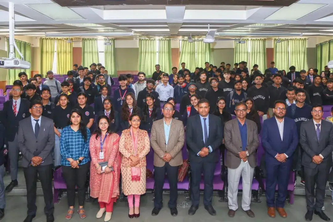 Group photo of students and faculty in an auditorium, front row in formal attire, green curtains in the background, audience seated behind them.