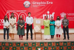 Eight people wearing masks stand in a line on a stage in front of a red backdrop with logos, a large cardboard box sits on a chair in the center.