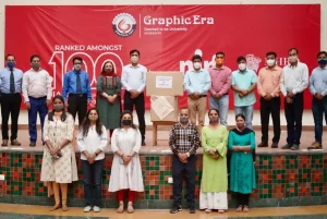 Group of people on a stage posing for a photo in front of a red Graphic Era banner, with a large cardboard box displayed on a stand at center.