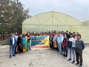 Group of people standing outdoors in front of a large greenhouse, holding a colorful banner for a community event.