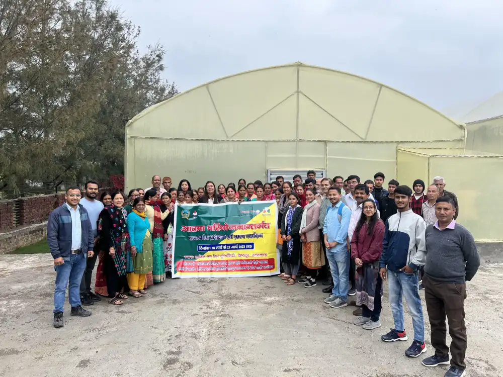 Group of people standing outdoors in front of a large greenhouse, holding a colorful banner for a community event.