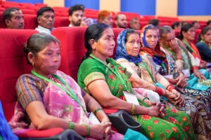 Group of Indian women in colorful sarees sitting in red theater seats at an event, listening attentively.
