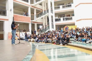 Female speaker on a stage addressing a large student audience in a multi-level campus atrium, using a microphone.
