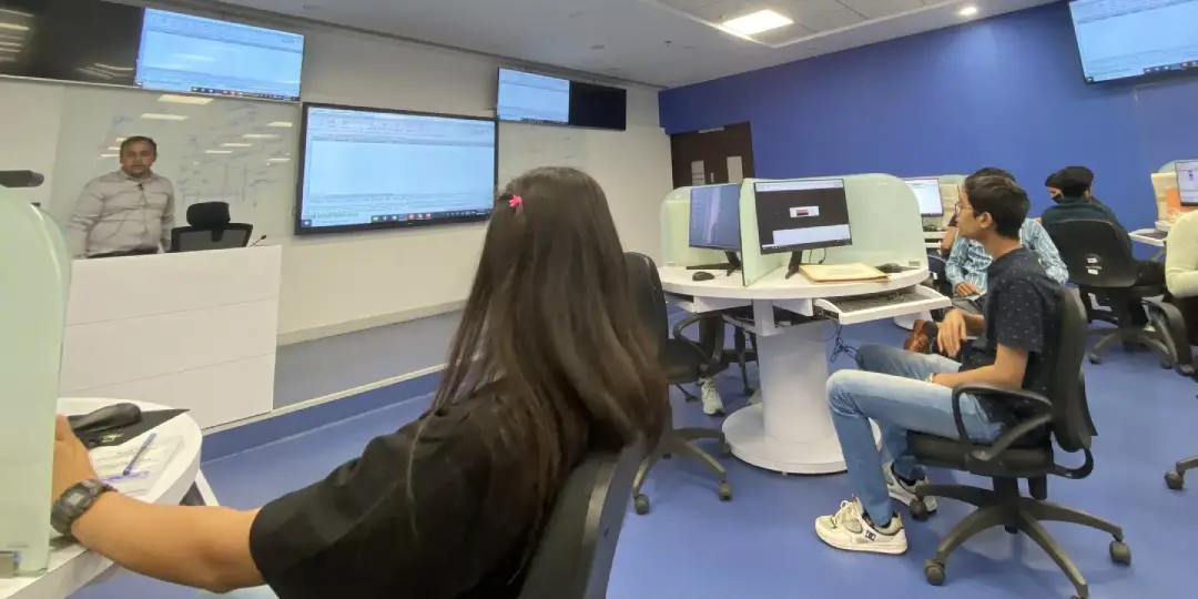 Instructor standing at front podium presenting to students in a computer lab with large wall screens behind himage shows multiple desks with computer stations and students seated at screens.