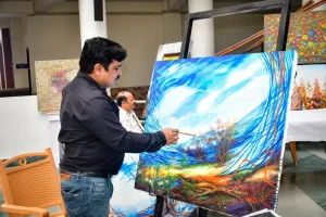 Artist painting a large, colorful landscape on an easel in an art studio/gallery.