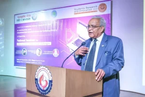 Older man in a blue suit speaks at a wooden podium with a microphone; a purple university banner is in the background.