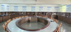 Large circular laboratory work area with an oval central table, brown cabinetry along the walls, and posters mounted on a white wall in the background.