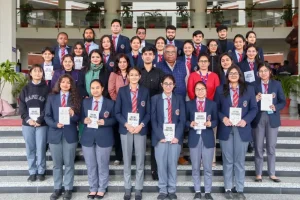 Group of students in blue blazers and red ties posing on school stairs with two adults at the back.