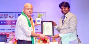 Bald man in white shirt and Indian flag scarf hands a framed certificate to a suited man on a stage with a potted plant beside them, during an award ceremony.